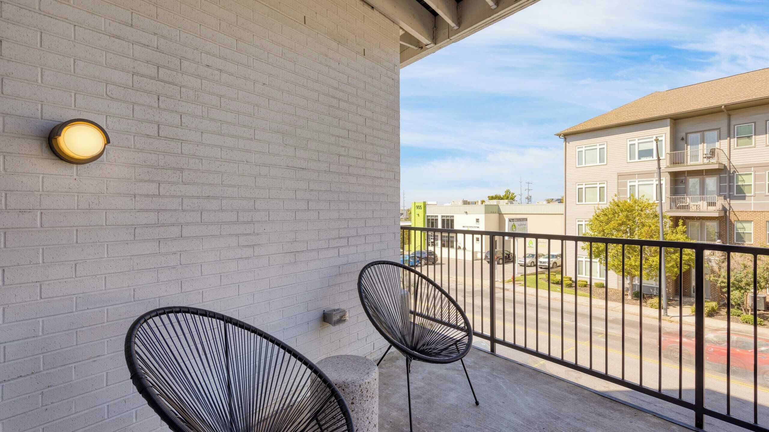 A balcony with two black chairs, a light fixture, and a view of a parking lot and buildings.