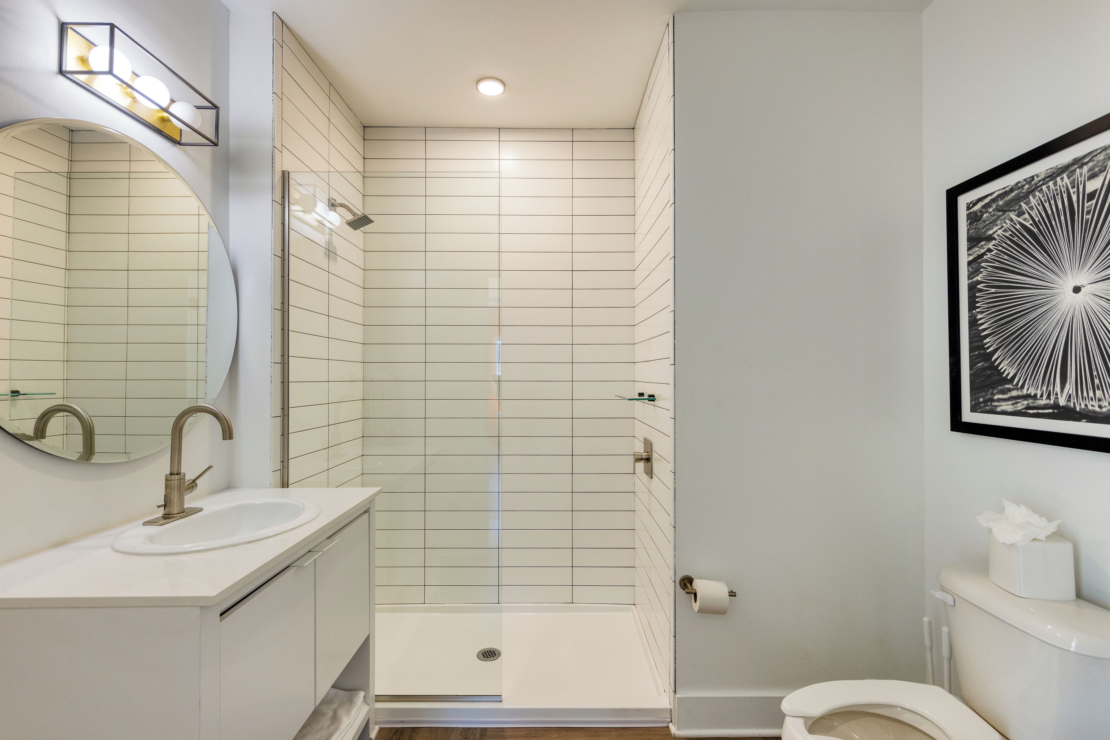 A modern bathroom with a sink, mirror, shower, toilet, and a framed artwork on the wall.