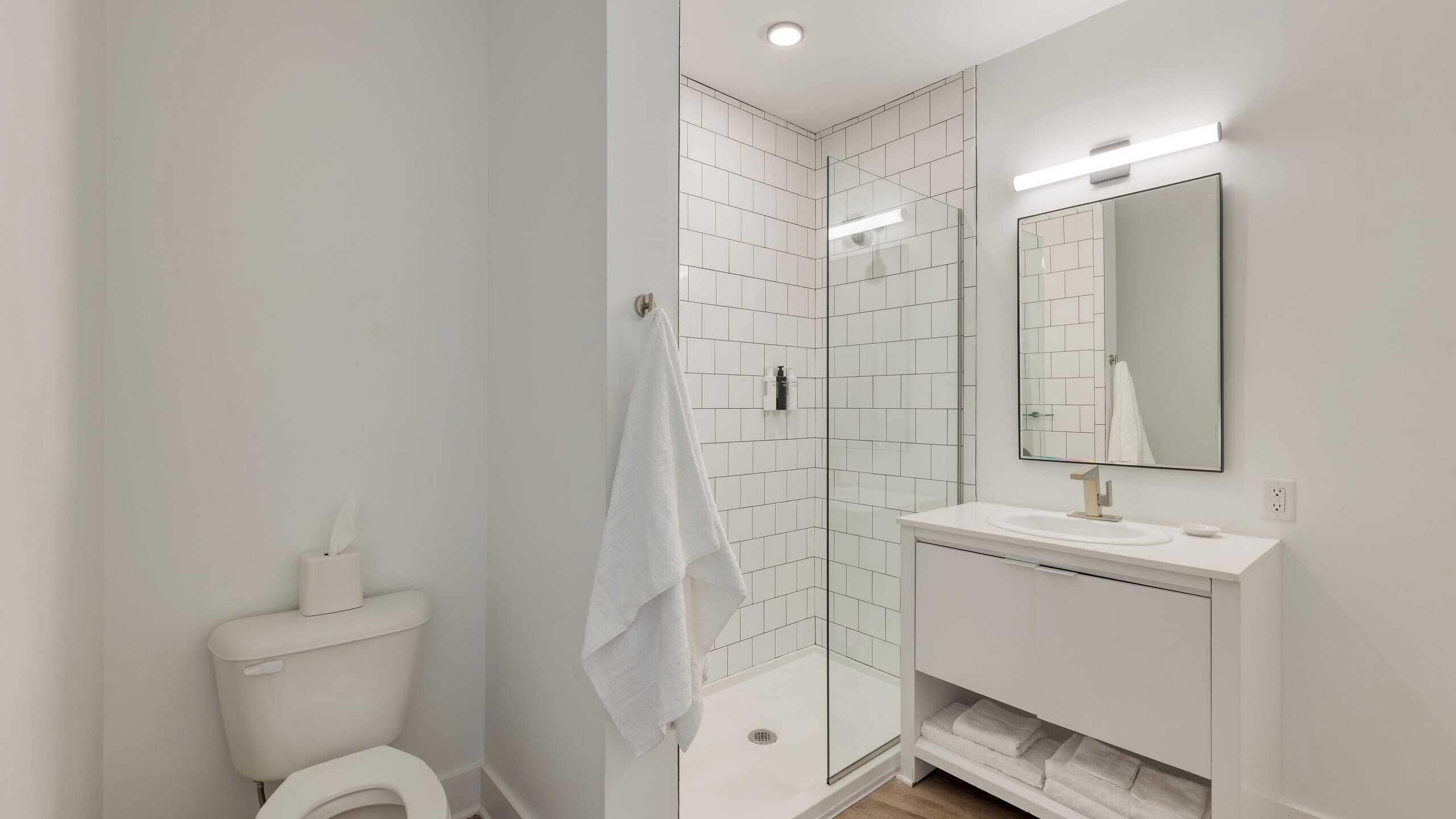 A modern bathroom with a glass shower enclosure, white tiles, a toilet, and a vanity with a mirror.