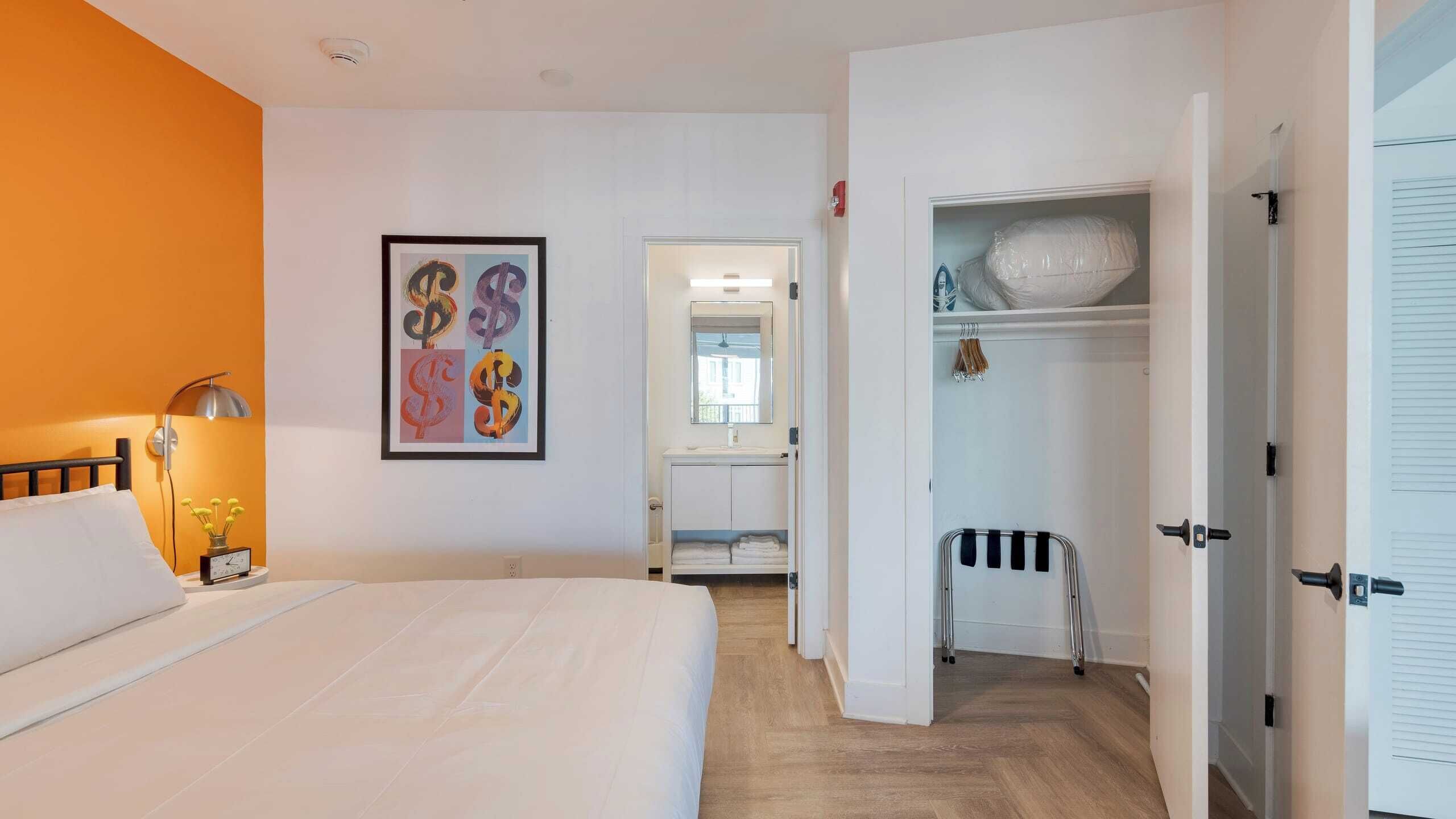Une chambre d’hôtel comprenant un lit, une table de chevet, une œuvre d’art encadrée, une armoire et une salle de bain visible à travers une porte ouverte.