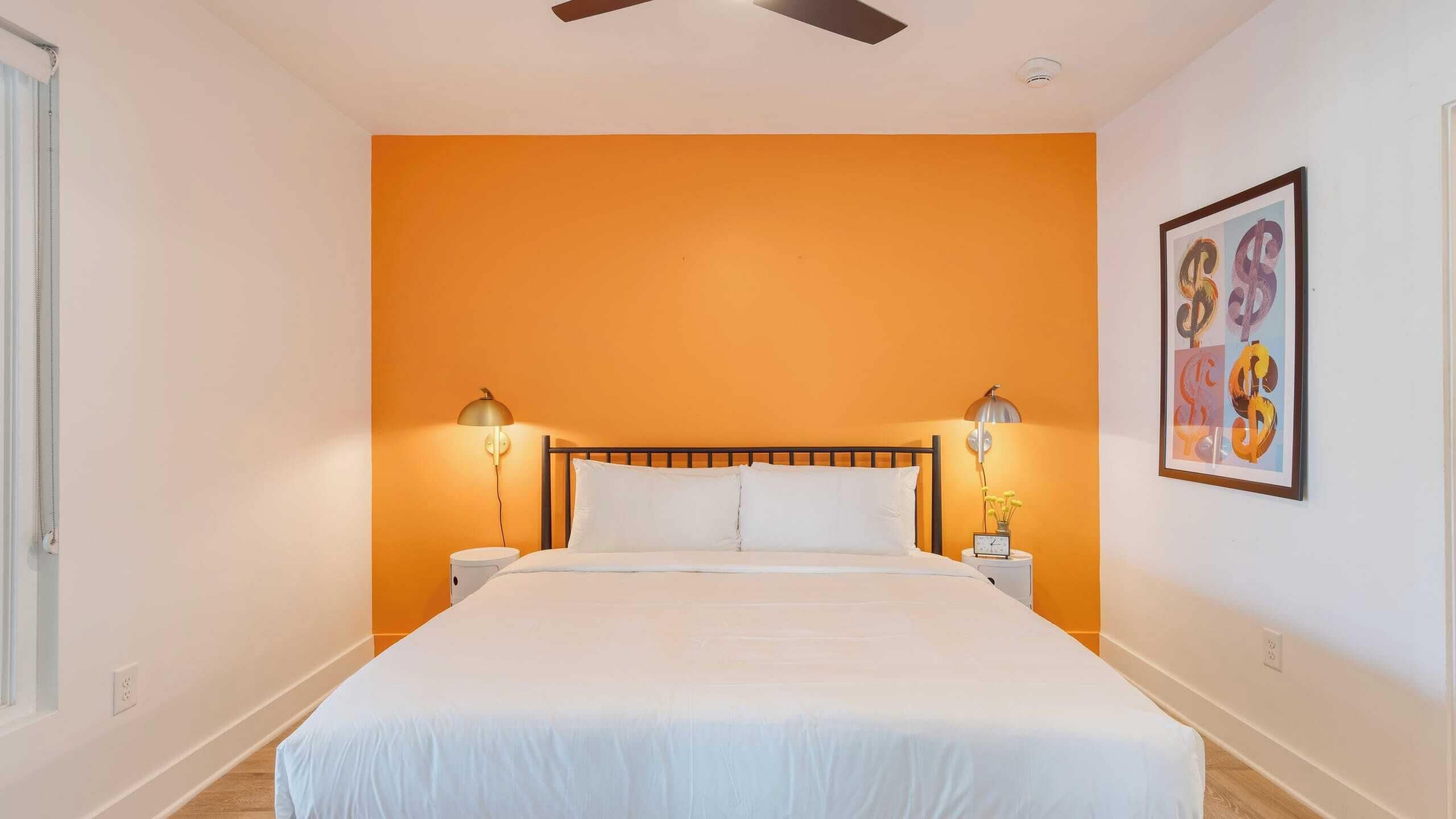 A bedroom with an orange accent wall, a bed with white bedding, two bedside lamps, and a framed artwork on the wall.