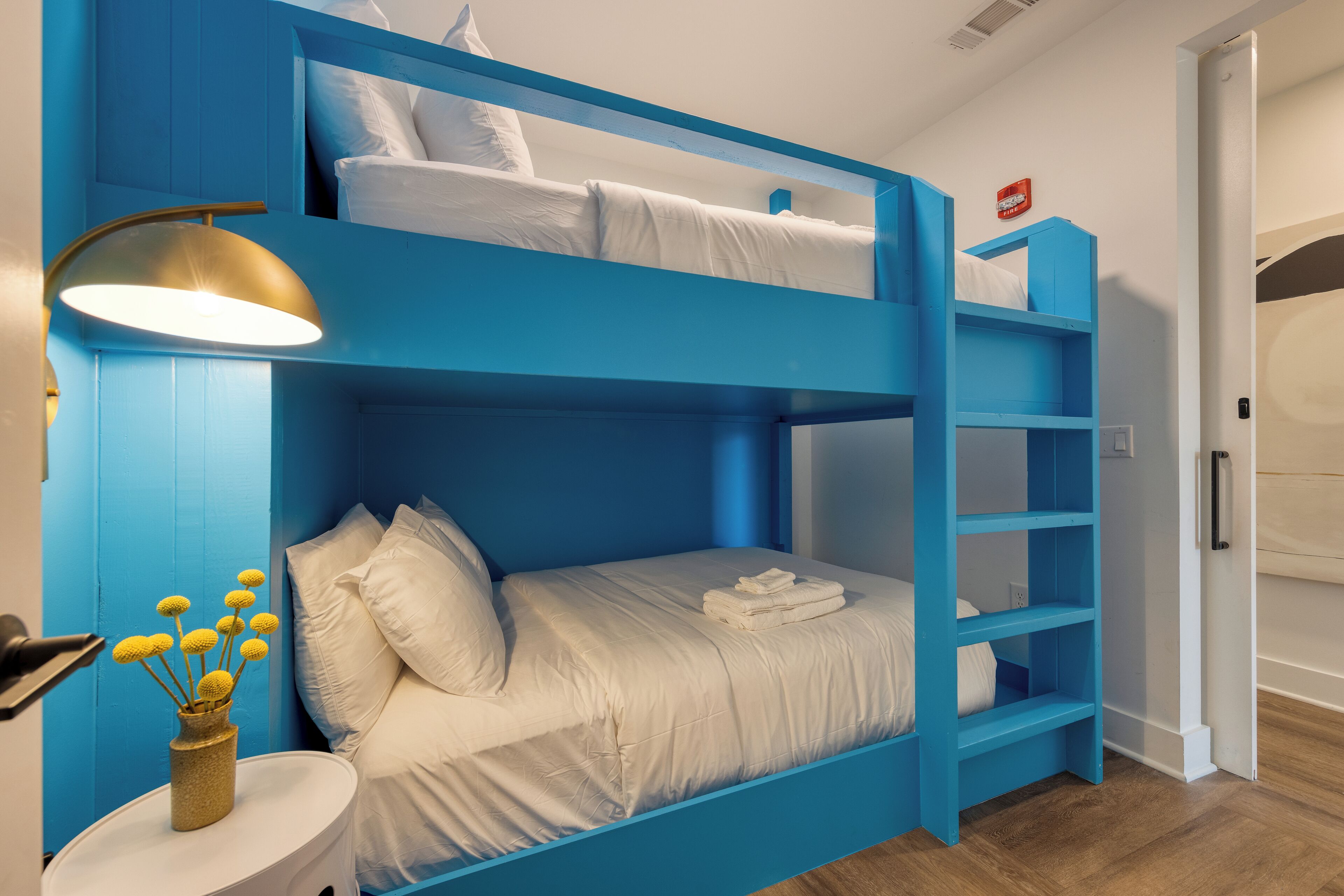 A bunk bed room with a blue color scheme, a bedside lamp, and a vase with yellow flowers.
