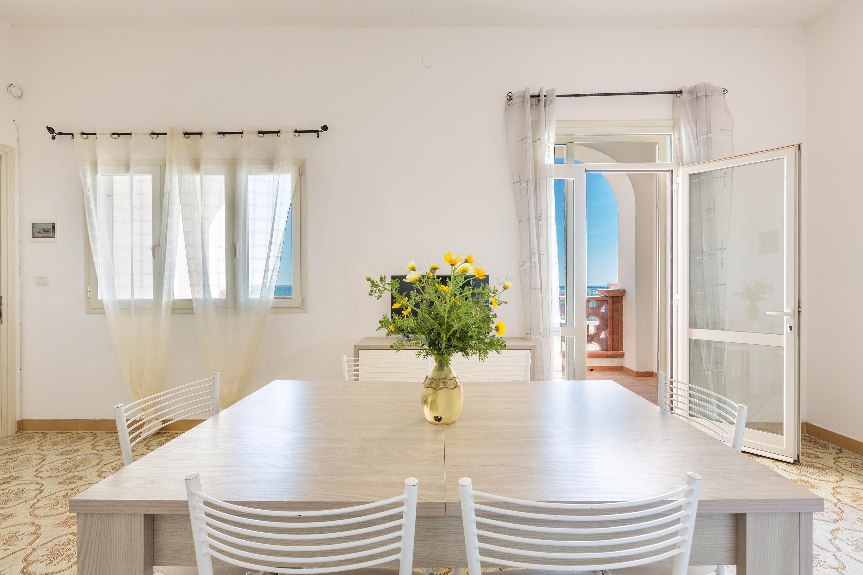 Un coin repas avec une table en bois, des chaises blanches, un vase rempli de fleurs jaunes et une vue sur l’extérieur grâce à une porte vitrée.