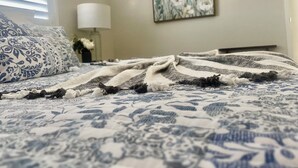 Een net opgemaakt bed met een geborduurde spreideken, een bedlampje en een bloemen schilderij aan de muur.