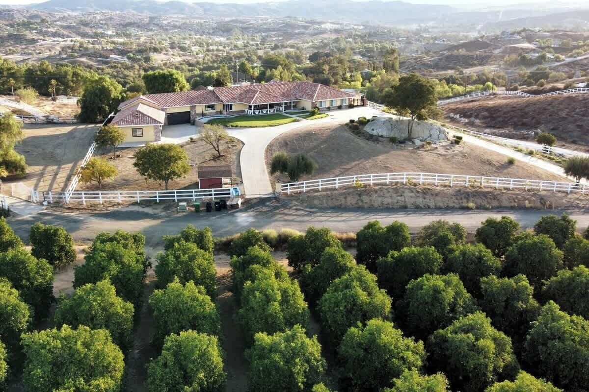 A large house with a red-tiled roof, surrounded by a white fence, and a driveway leading to a gated entrance.