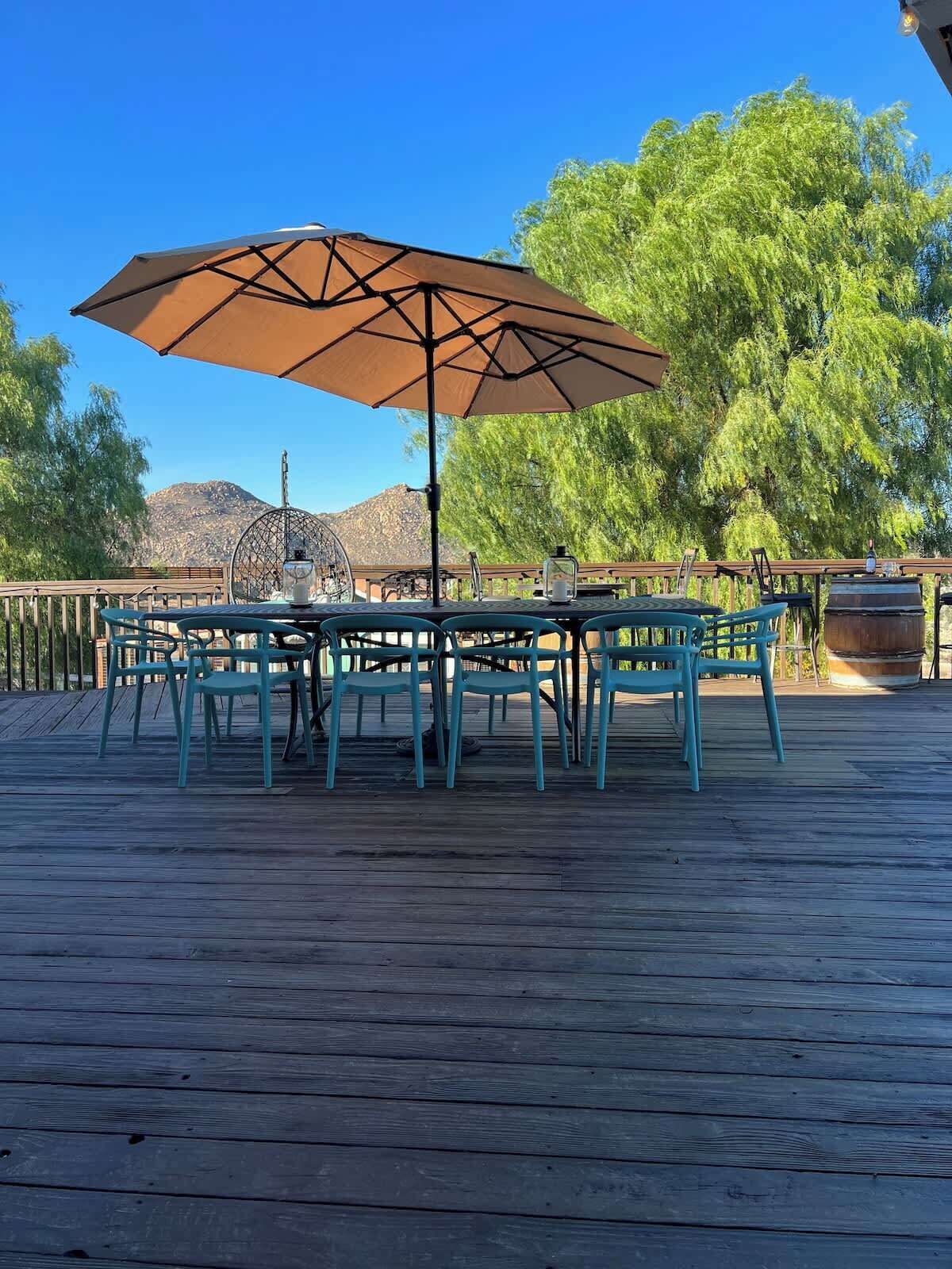 A wooden deck with a table and chairs under a large umbrella, surrounded by trees and a mountain in the background.