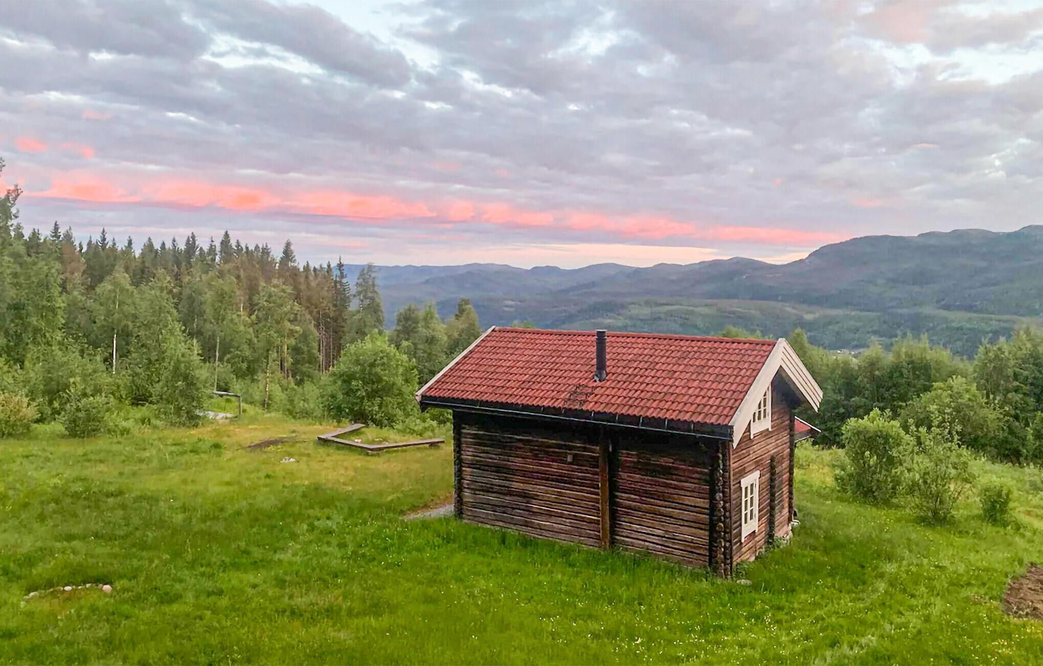 A rustic cabin with a red roof, surrounded by lush greenery and mountains in the background.