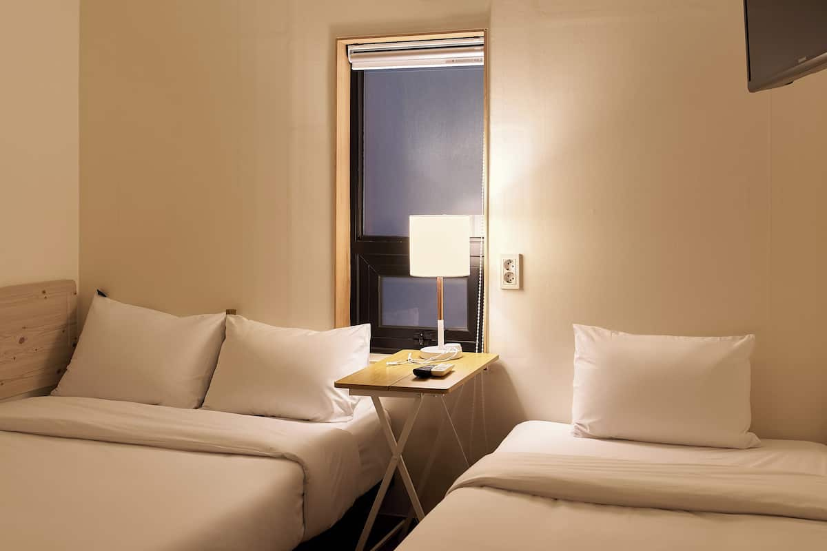 A hotel room with two beds, a bedside table with a lamp, and a window with blinds.