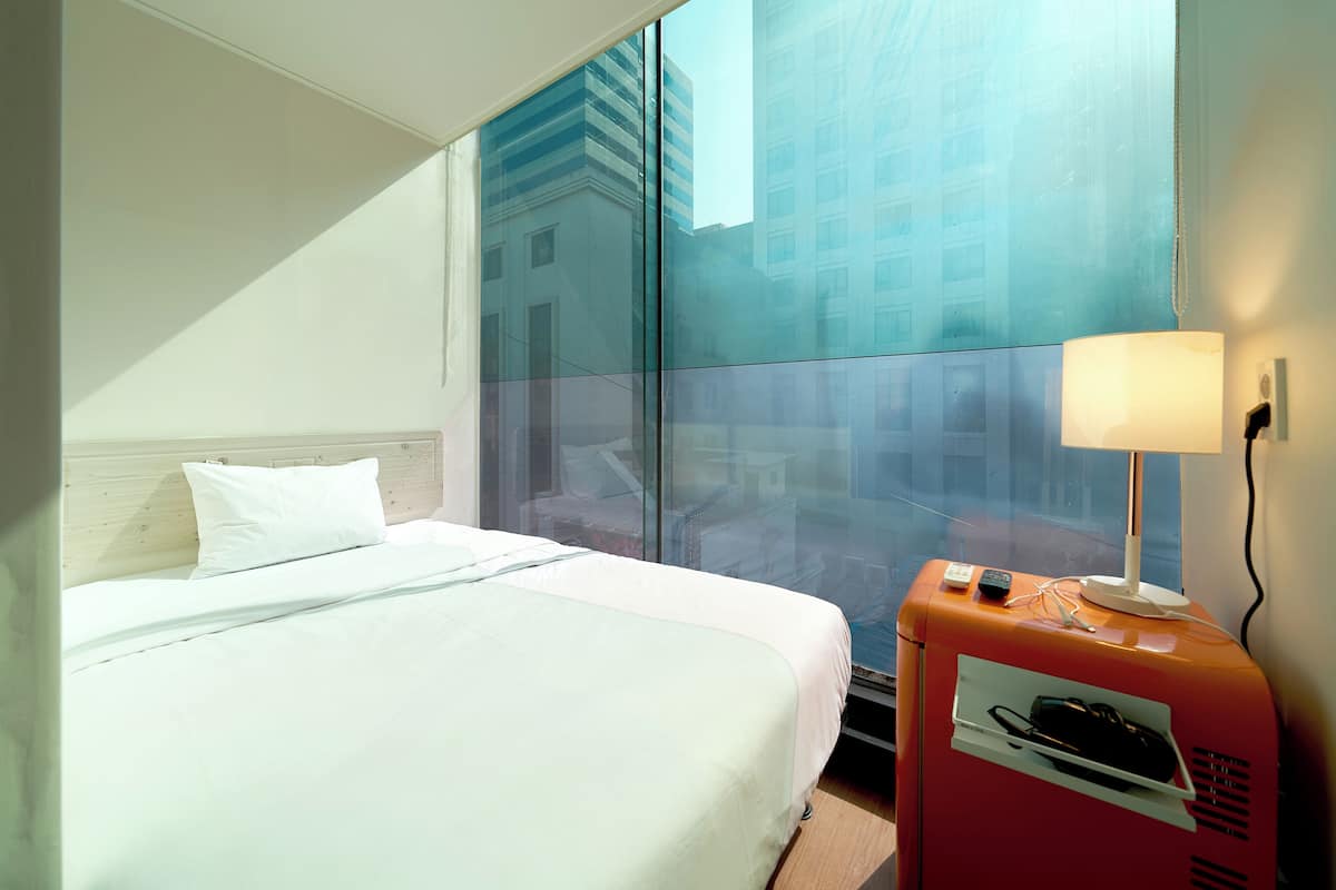 A hotel room with a bed, a nightstand with a lamp, and a view of buildings through a large window.