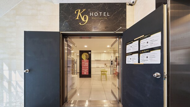 The entrance to a hotel with a 'Welcome' mat, a reception desk, and a sign indicating a 3PM check-in policy. - K9 Myeongdong Seoul (Seoul)