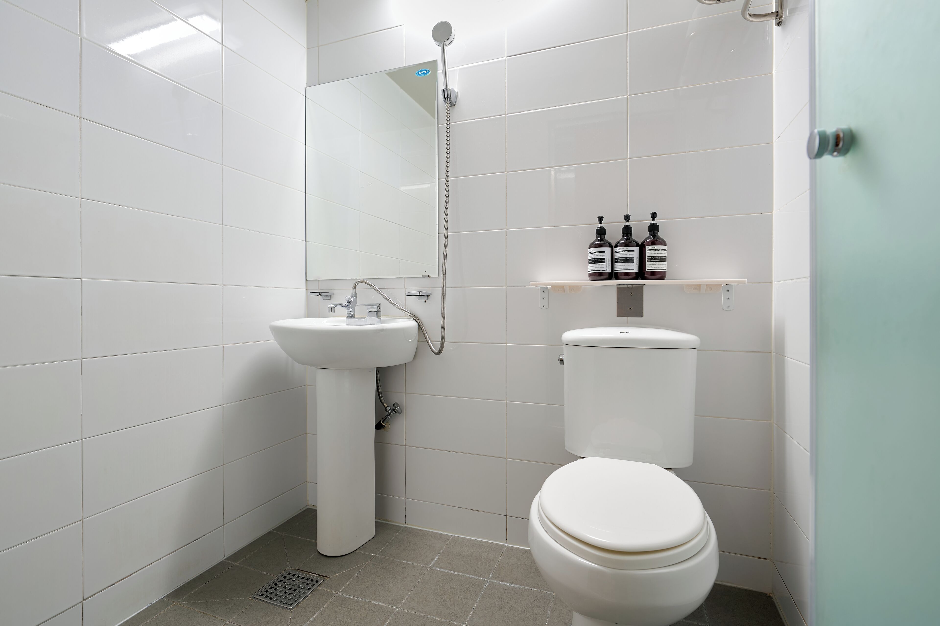 Ein Badezimmer mit weißem Waschbecken, WC und Dusche. Über dem WC befindet sich ein Regal mit zwei Flaschen.