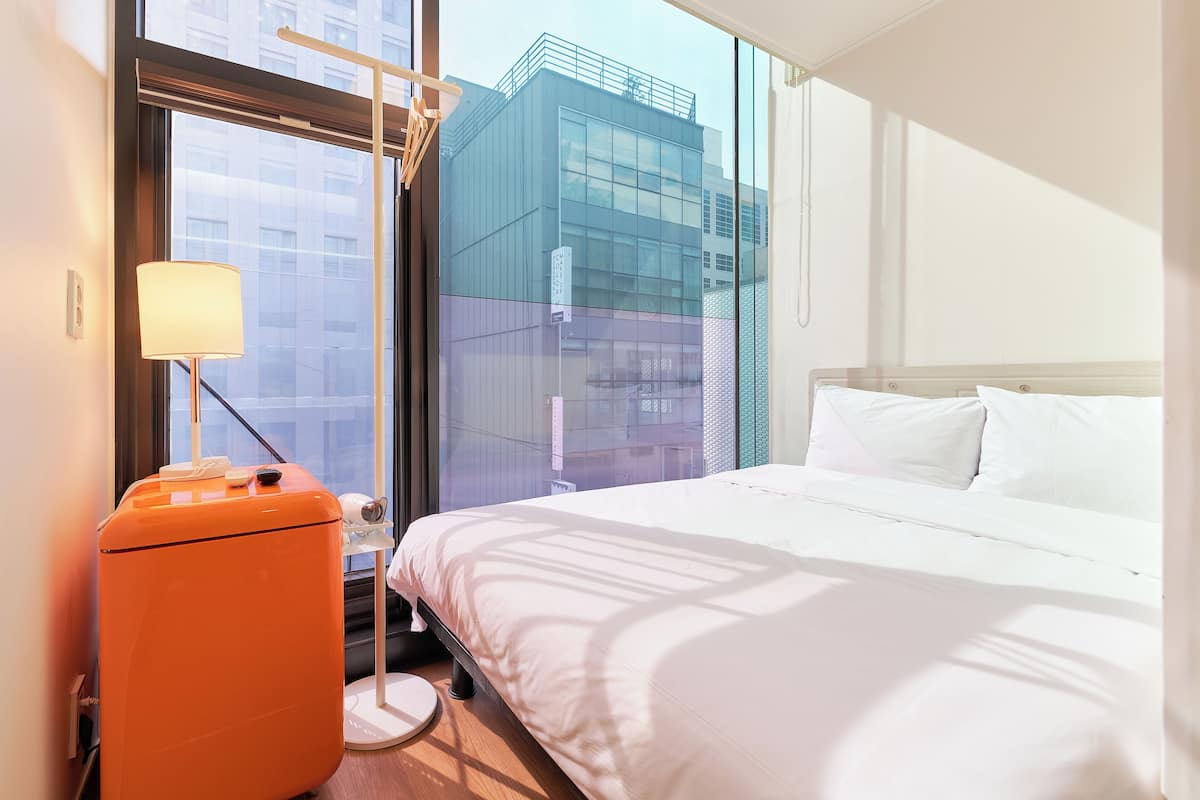 A hotel room with a bed, a lamp, a suitcase, and a window with a view of a modern building.