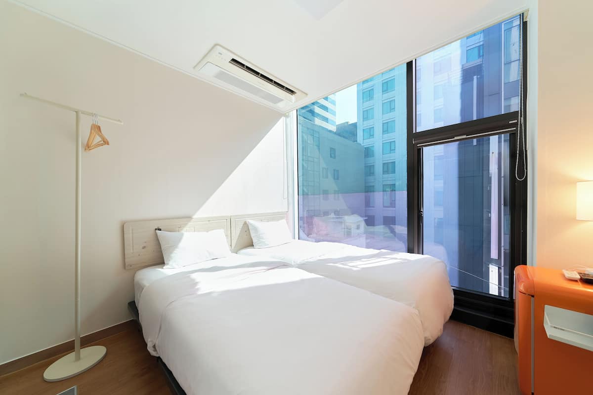 A hotel room with two beds, a standing heater, a window with a view of buildings, and a bedside table with a lamp.