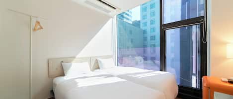 Une chambre d’hôtel avec deux lits, un radiateur mobile, une fenêtre donnant sur des immeubles et une table de chevet avec une lampe.