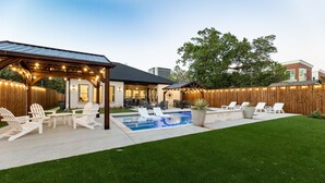 Un patio trasero con piscina, sillas de descanso y una pérgola con luces colgantes.