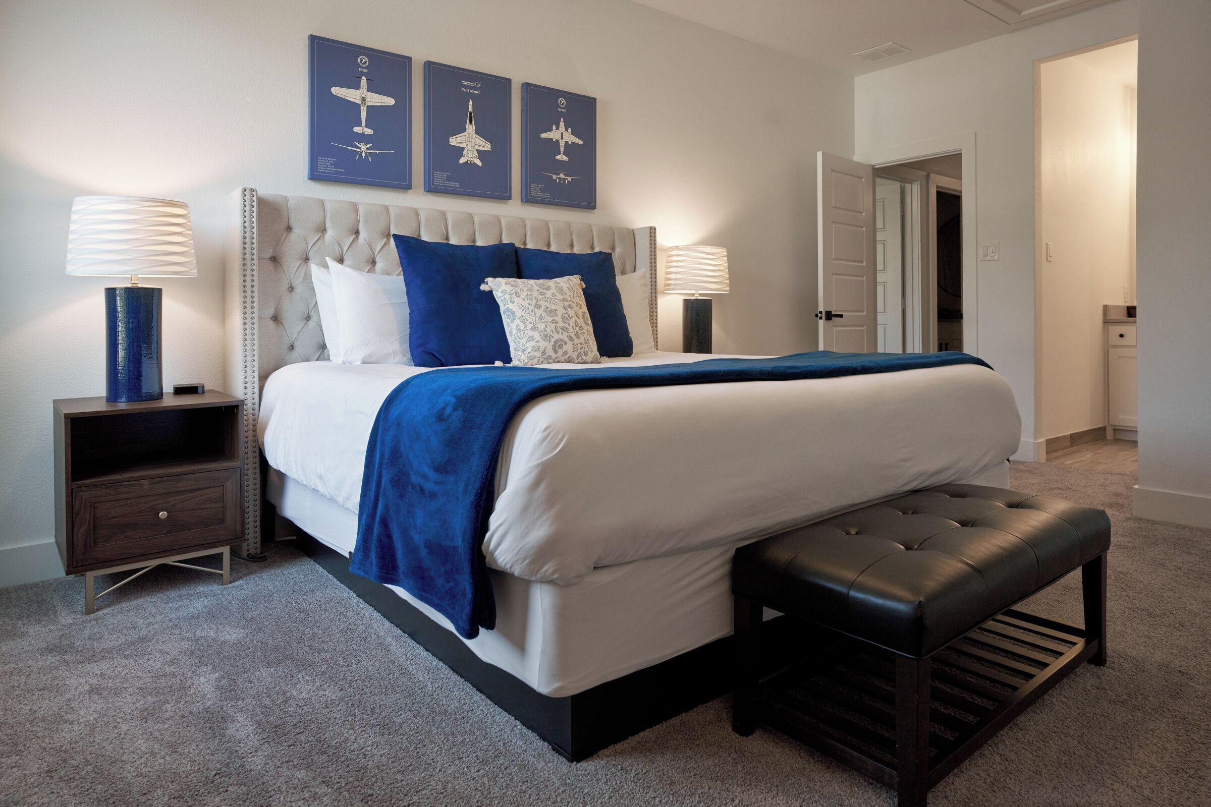 A bedroom with a tufted headboard, a large bed with white and blue bedding, two bedside tables with lamps, and a bench at the foot of the bed.