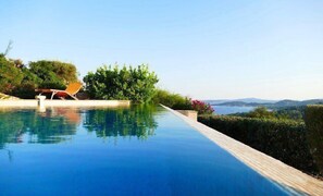 A clear blue infinity pool with a wooden deck, surrounded by lush greenery and a view of distant hills. - Villa with pool for 10 people (LES ISSAMBRES)