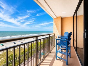Un balcon avec vue sur la plage et l’océan, deux chaises bleues et une porte vitrée.