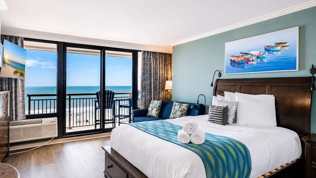 Een hotelkamer met een groot bed, uitzicht op het strand, een balkon met stoelen en een schilderij aan de wand.