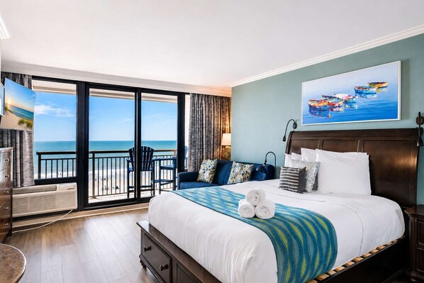 Une chambre d’hôtel avec un grand lit, une vue sur la plage, un balcon avec des chaises et un tableau au mur.