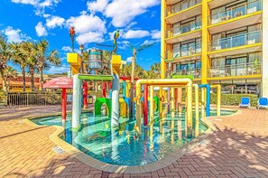 A swimming pool with colorful play structures, a multi-story building with balconies, and palm trees in the background.
