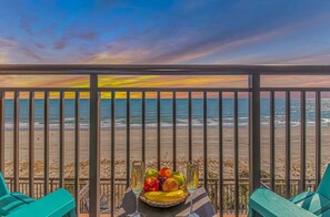 Ein Balkon mit Blick auf Strand und Meer, ein Tisch mit Früchten und Gläsern sowie türkisfarbene Stühle.