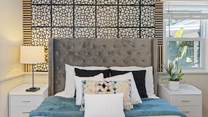 A neatly arranged bedroom with a tufted headboard, a variety of pillows, a bedside lamp, and a window with a view of greenery. - Old Town Monrovia Hot Tub Retreat (Monrovia)