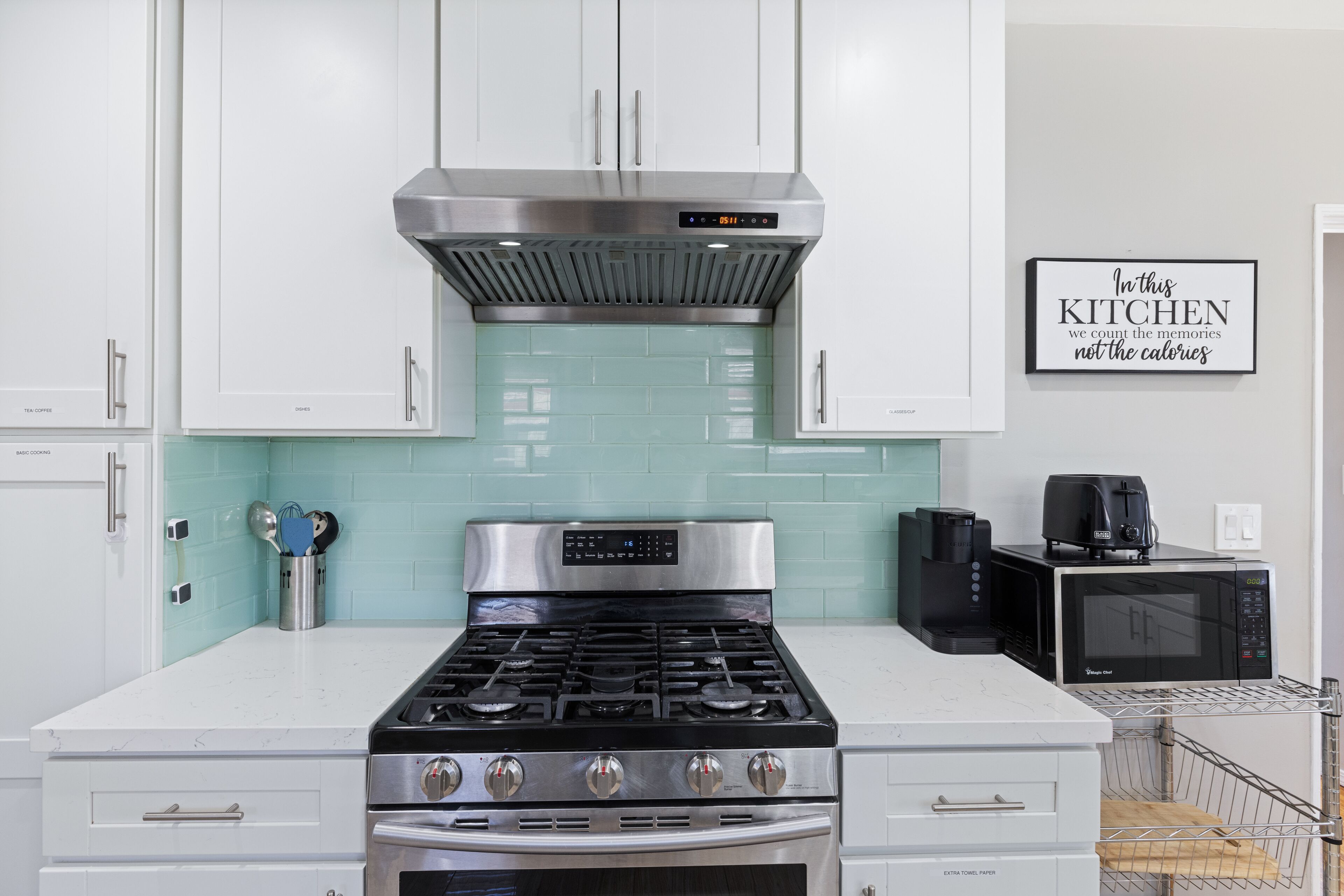 A modern kitchen with white cabinets, a stainless steel range hood, a stove, an oven, a microwave, and a coffee maker.