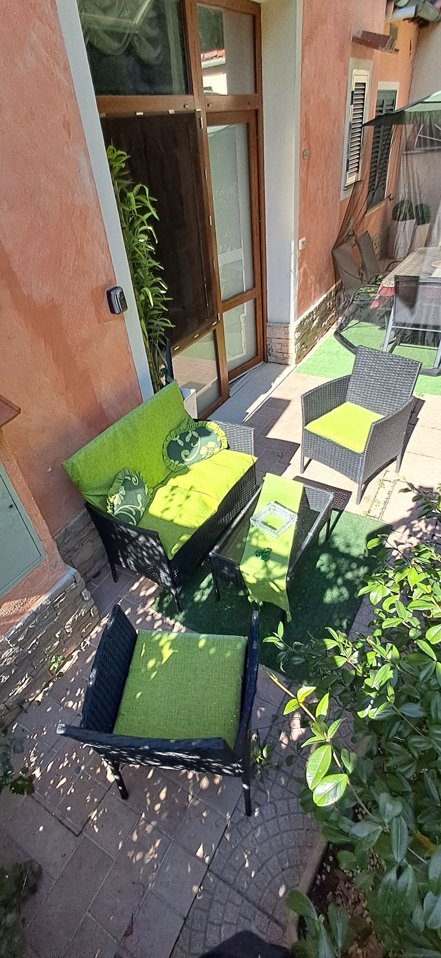 Un espace extérieur aménagé avec des meubles en osier et des coussins verts, une entrée avec une porte en verre et un petit jardin avec des plantes.