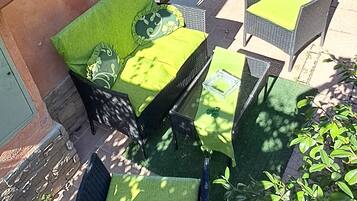Un espace extérieur aménagé avec des meubles en osier et des coussins verts, une entrée avec une porte en verre et un petit jardin avec des plantes.