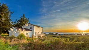 A white house with a deck, surrounded by grass and trees, overlooking a body of water at sunset. - Cozy Oceanview Hideaway#2 on Golf Course/Airport (Whitethorn)