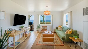 A living room with a sofa, coffee table, and TV, a wooden table with a plant, and a view of the outdoors. - Cozy Oceanview Hideaway#2 on Golf Course/Airport (Whitethorn)