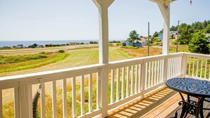 A wooden deck with a white railing, a round metal table, and a view of a coastal landscape. - Cozy Oceanview Hideaway#2 on Golf Course/Airport (Whitethorn)