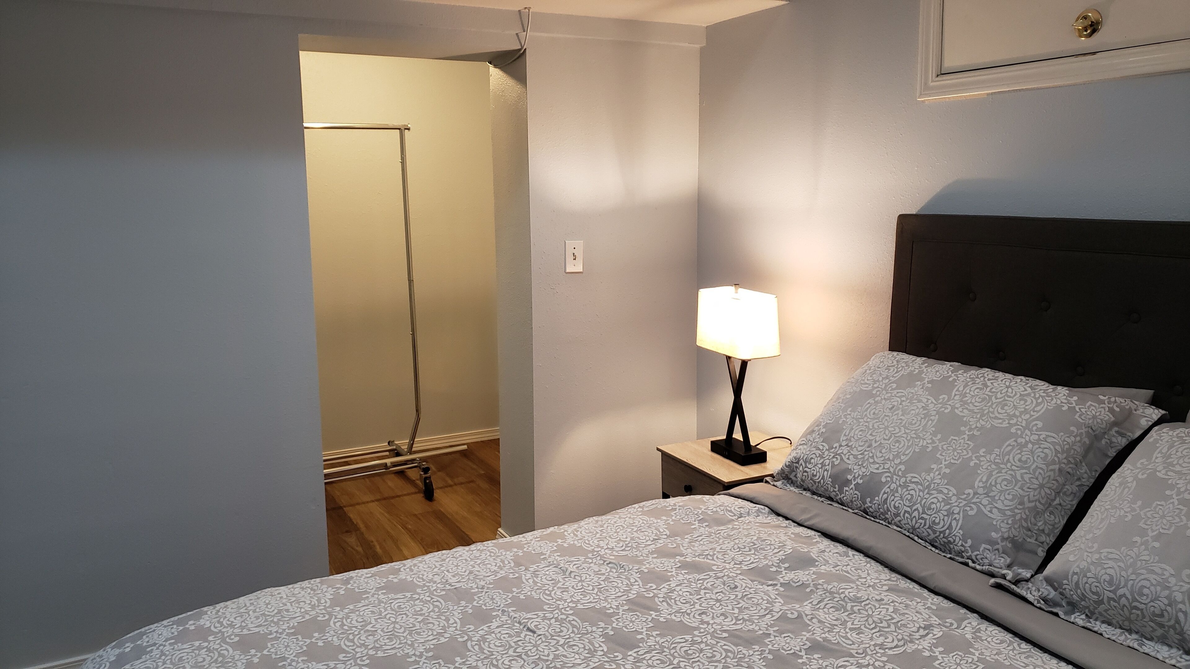 A bedroom with a bed, a nightstand with a lamp, a dark headboard, and a door leading to another room.