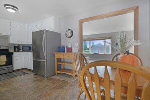 A compact kitchen with white cabinets, a stainless steel refrigerator, a microwave, and a coffee maker. There's a wooden table with chairs and a vase with a plant. A clock and a window with a view of a house are also visible.