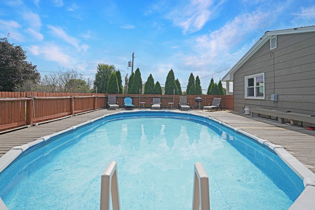A rectangular swimming pool with a wooden deck and lounge chairs, surrounded by a wooden fence and trees.
