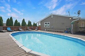 A swimming pool with a wooden deck, surrounded by lounge chairs and a house with satellite dishes.