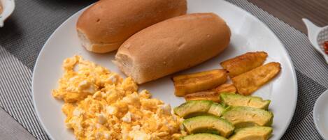 A plate with scrambled eggs, two hot dogs, fried plantains, and sliced avocado.
