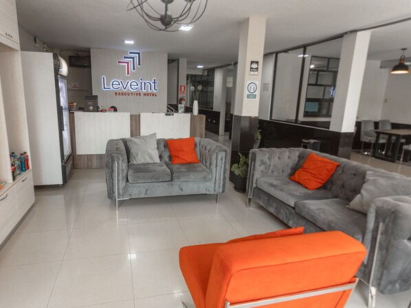Un moderno vestíbulo de hotel con dos sofás grises, uno con un cojín naranja, y una silla naranja brillante. Se ve un cartel del Hotel Ejecutivo Leveint.