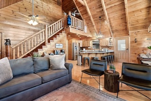 A spacious living area with a gray sofa, a wooden staircase, and a kitchen in the background.