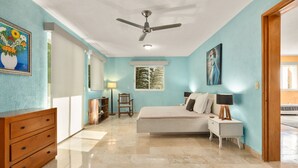 A bedroom with a bed, a dresser, a ceiling fan, a window with a view, and a painting on the wall.