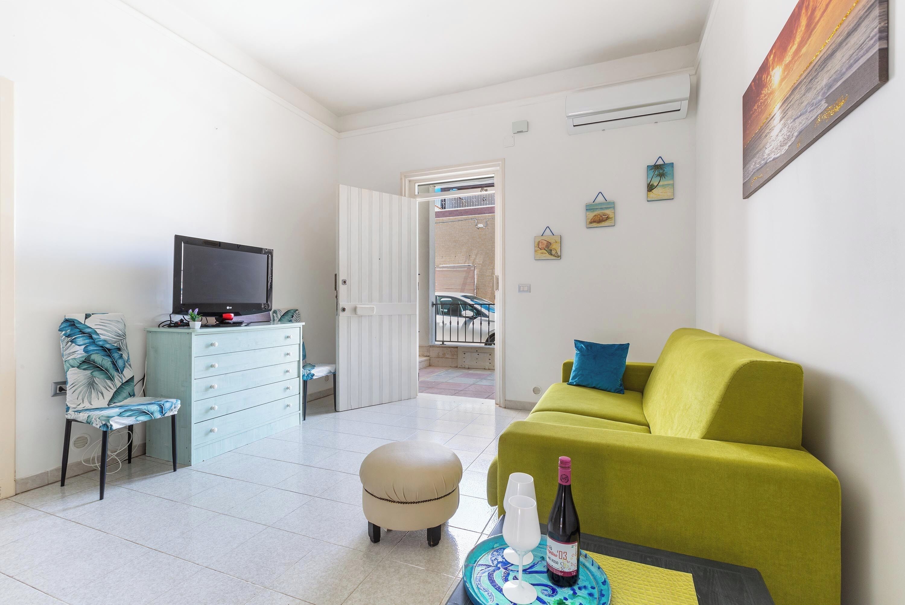 Una sala de estar moderna con un sofá verde, un pequeño taburete redondo, un televisor sobre un mueble bajo y una mesa de comedor con una botella y vasos.