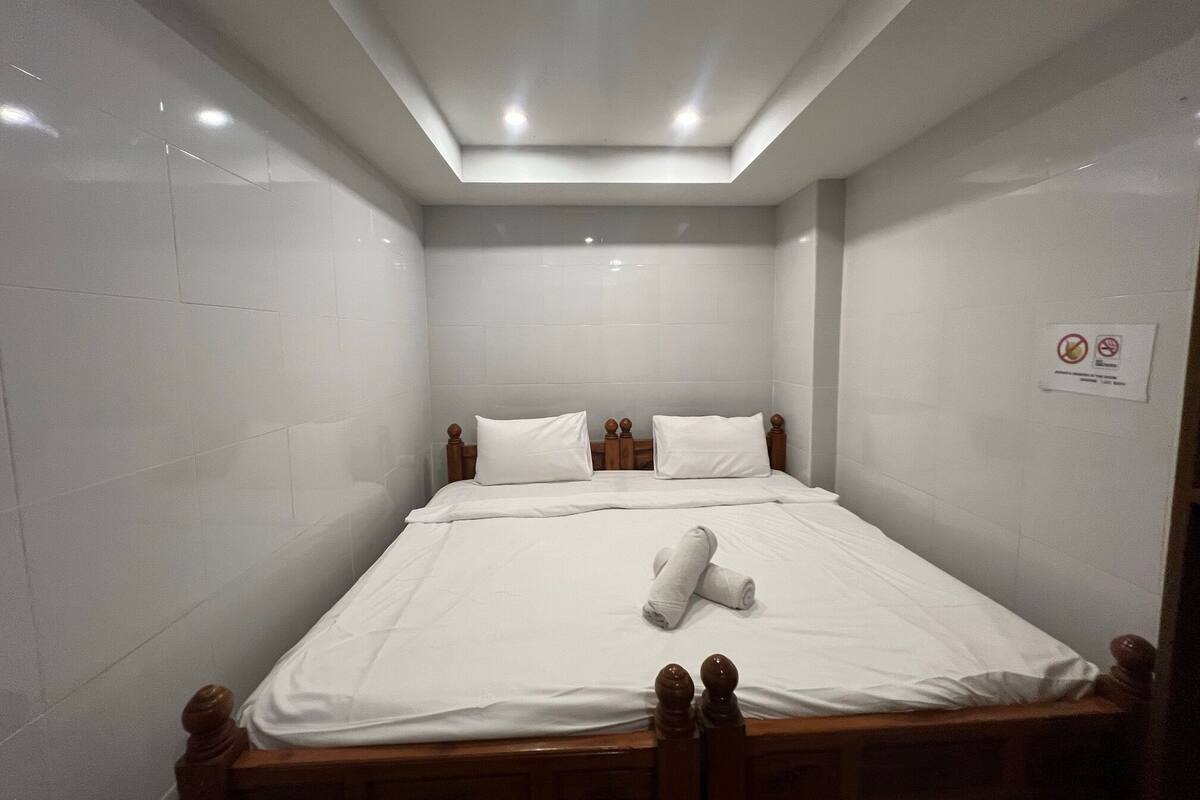 Ein kleiner, gut beleuchteter Raum mit einem Einzelbett, weißer Bettwäsche und einem Holzkopfstück.