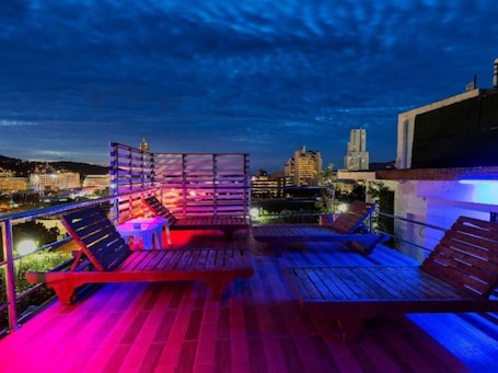 A rooftop terrace with wooden loungers and a small table, overlooking a cityscape at night.