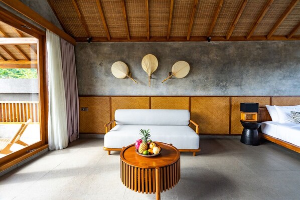 Une chambre moderne avec un canapé, une table basse ronde en bois sur laquelle sont disposés des fruits, et un lit avec une table de chevet.