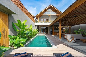 A modern pool area with a wooden deck, lounge chairs, and a covered seating area with a table and chairs. - Jiwoku Villas (Canggu)