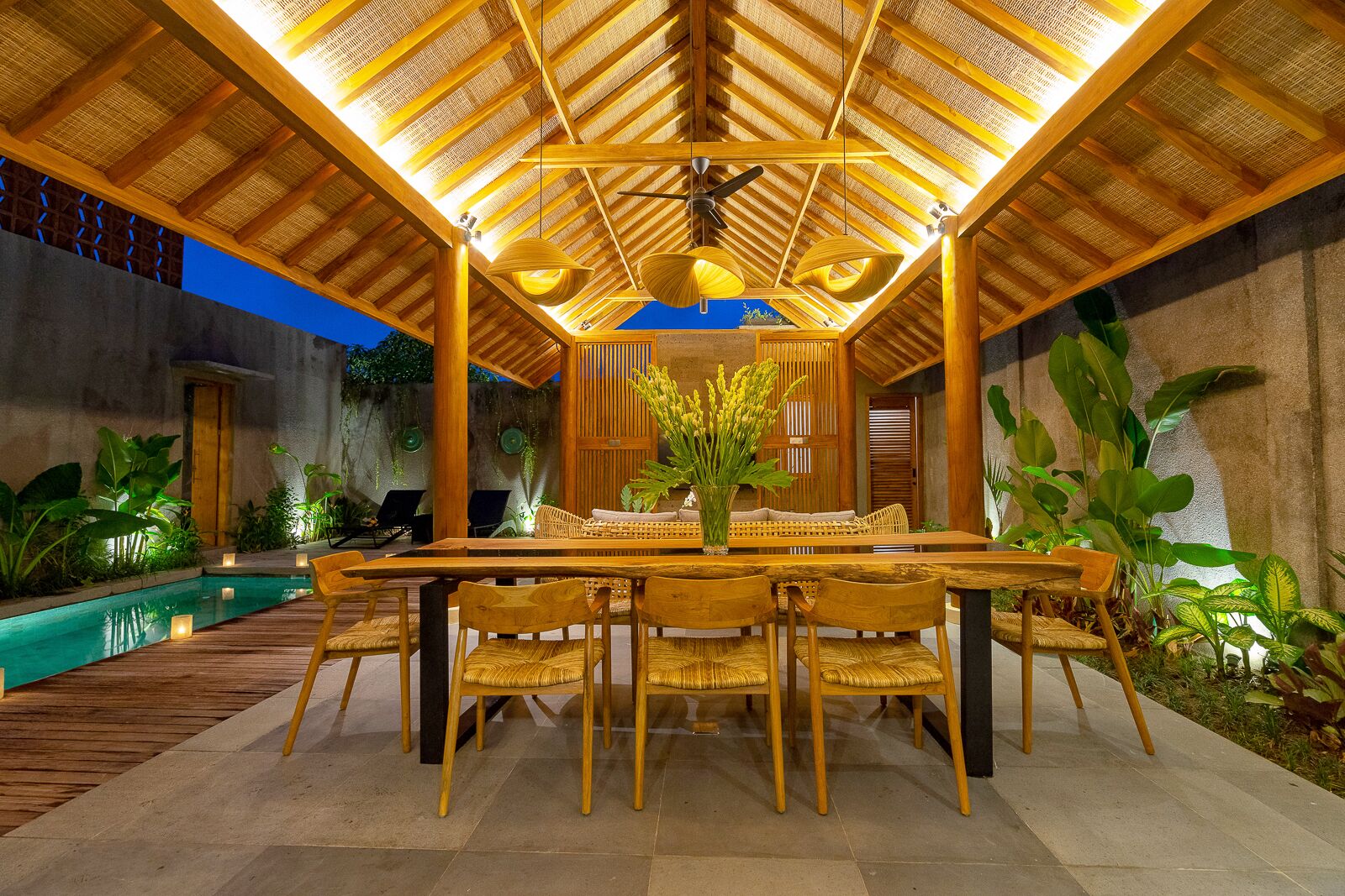 An outdoor dining area with a wooden table and chairs, surrounded by plants and a pool.