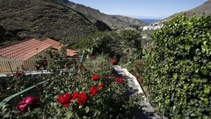 Un paisaje montañoso con vista al mar, un edificio con techo de tejas rojas y un jardín con flores rojas.