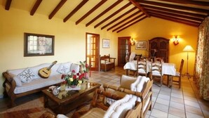 Un salón acogedor con techo de madera, un sofá con cojines decorativos, una mesa de centro con un jarrón de flores y una zona de comedor con una mesa y sillas.