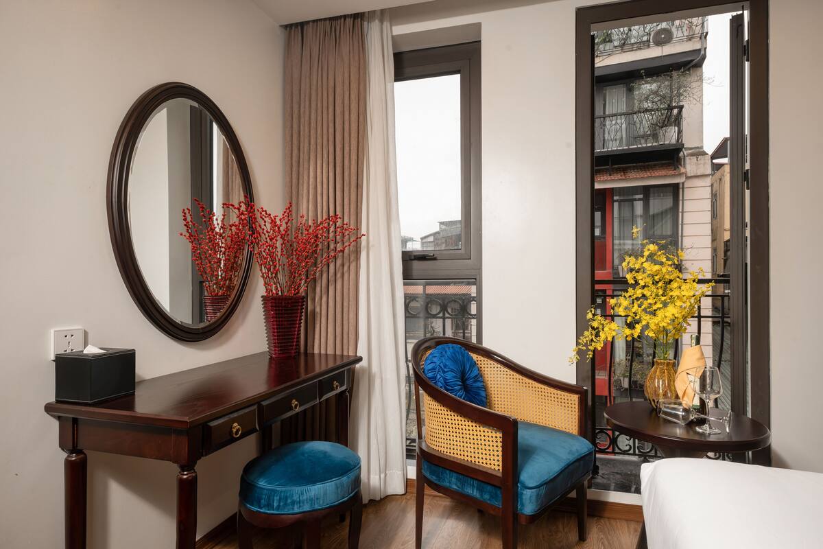 A hotel room with a wooden desk, a chair with blue cushion, a bed, a mirror, and a window with a view of buildings.